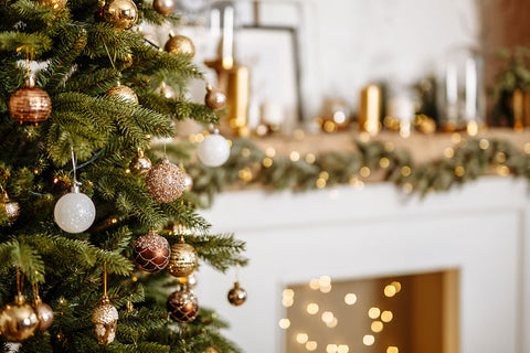 A close-up of a decorated Christmas tree with green branches, gold and white ornaments, and warm string lights, set in front of a blurred fireplace mantel with holiday decor.