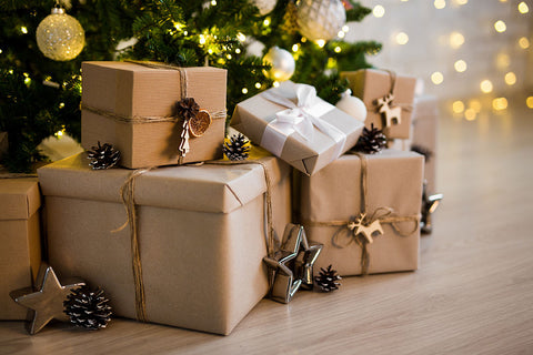 A collection of Christmas wrapped gift boxes and packages under a decorated tree. The packages are wrapped in natural brown kraft paper and white paper, tied with twine, and decorated with pinecones, metallic star ornaments, and wooden reindeer cutouts.