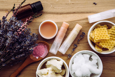 A styled flat-lay of natural lip balm ingredients on a wooden surface, including two lip balm tubes, a small amber bottle, bowls of beeswax, white coconut oil, pink powder, and dried lavender flowers.