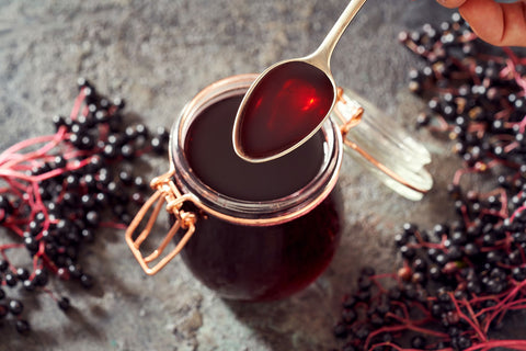 A glass jar filled with dark red elderberry syrup, with a spoon lifting some of the syrup. Fresh elderberry berries with pinkish stems are scattered around on a textured gray surface.