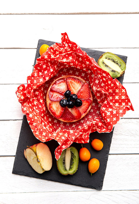 A round fruit tart or pastry topped with sliced strawberries and blueberries, wrapped in red-and-white patterned paper and presented on a black slate board with sliced kiwi, apple, and small orange fruits on a white wood background.